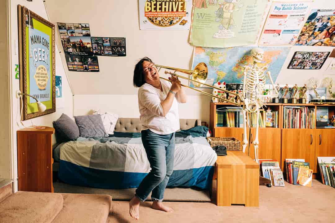 A barefoot woman plays the trombone in a creatively-decorated room.