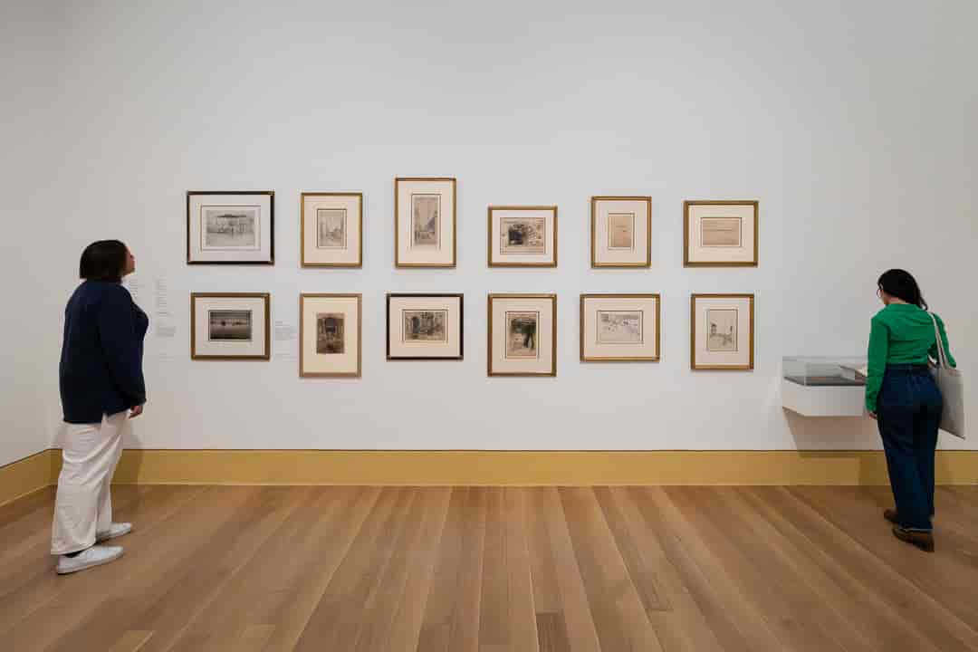 An image of paintings on the wall at a museum, with two people looking at them.