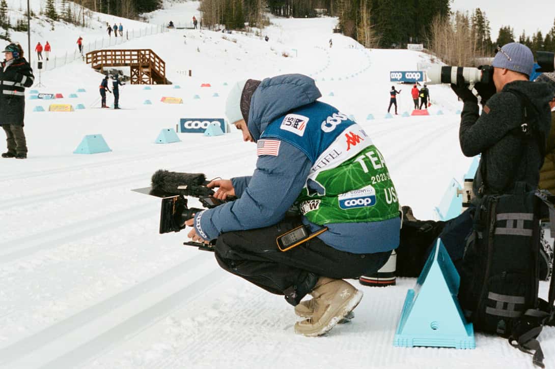 A person holding a camera crouches on snow during a ski race.