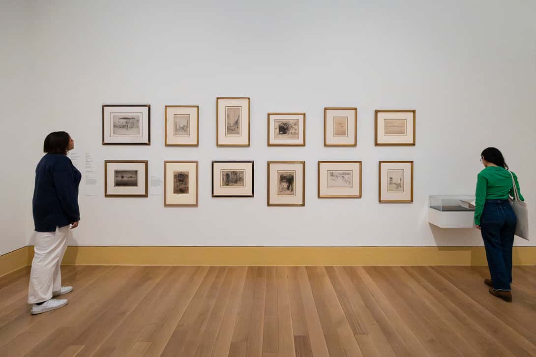 An image of paintings on the wall at a museum, with two people looking at them.