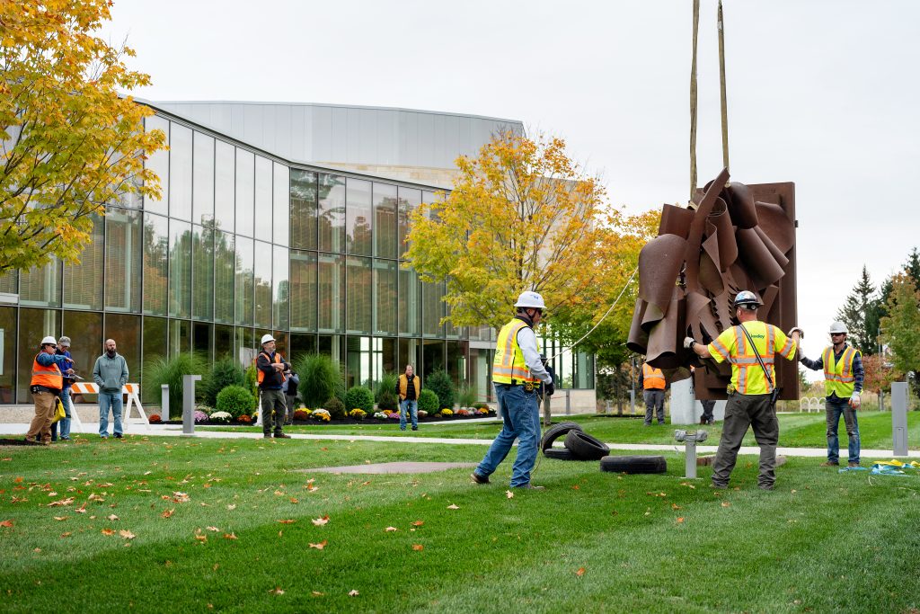 Workers in hardhats directing the placement of a metal sculpture putdoors.