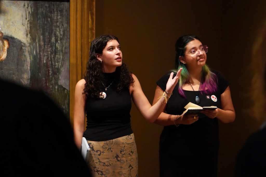 A woman holding a notebook speaks in front of a painting in a museum.