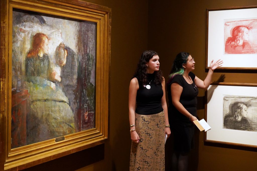 two woman standing in front of artwork in a museum. One woman points to a hanging painting to her left as she speaks.