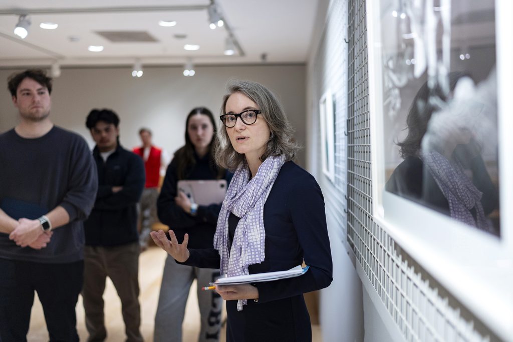A woman holding a notebook speaks to students in front of an art piece.
