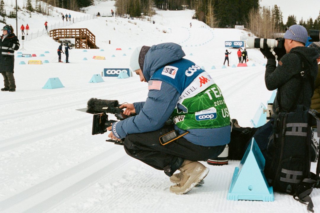 A person holding a camera crouches on snow during a ski race.
