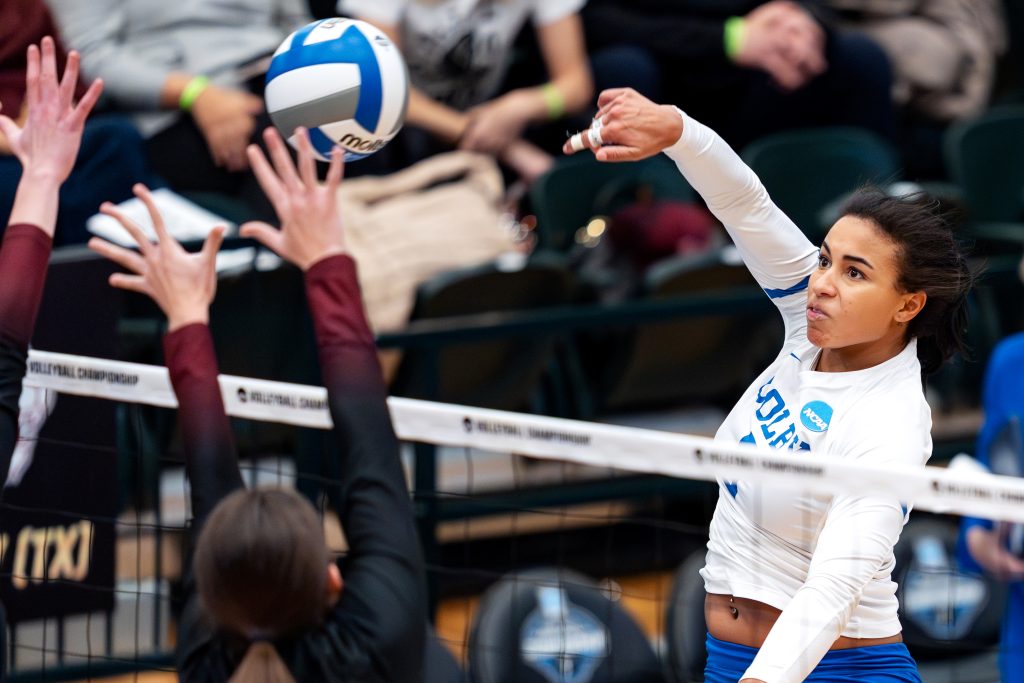 A volleyball player hits the ball over the net as two rival players attempt to block with arms outstretched.
