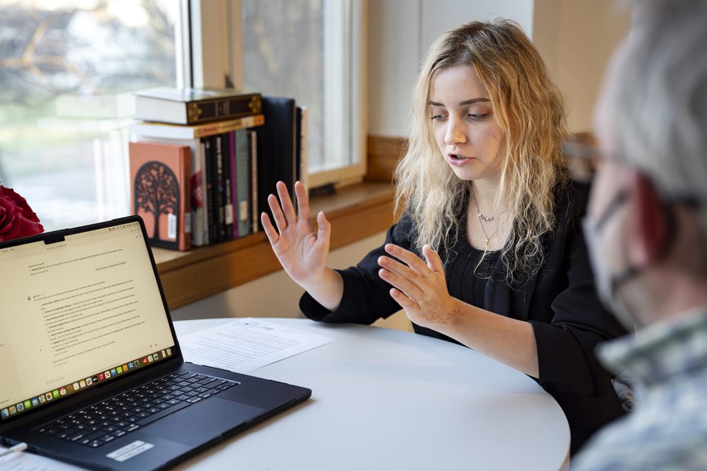 A sitting woman raises her hands while talking to a man seated at a table with an open laptop on it.