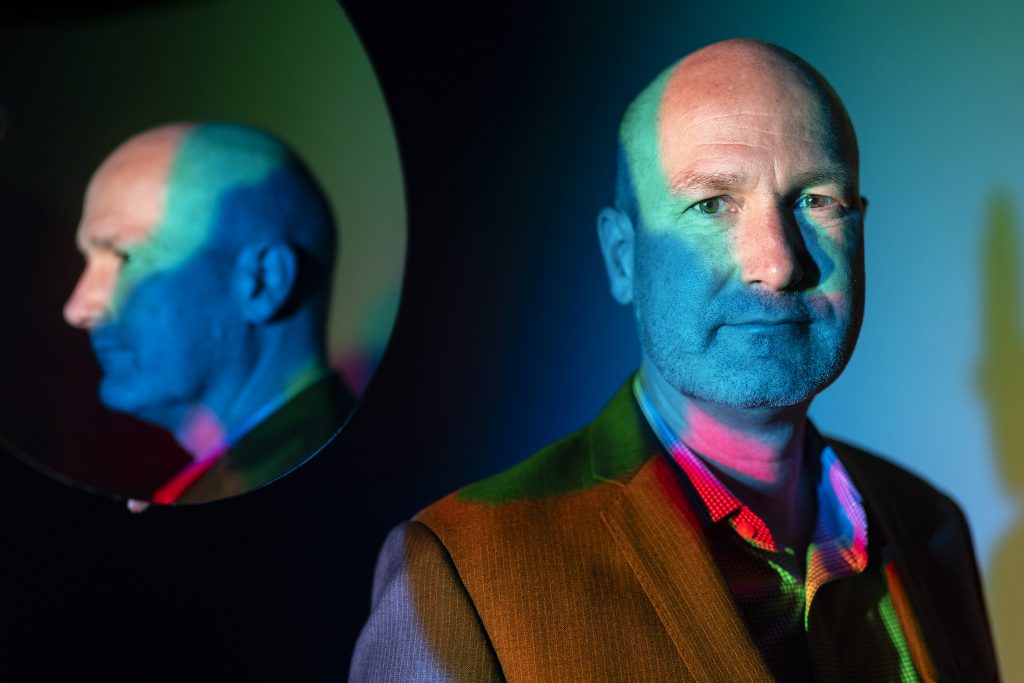 A man poses for a photo with a mirror behind him showing his reflection.