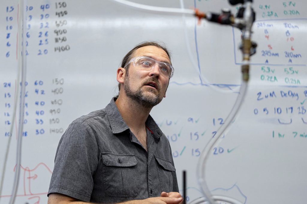 A professor wearing safety glasses in front of a whiteboard looks forwrd.