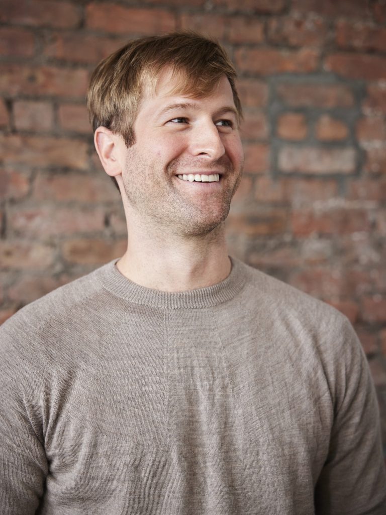 A man wearing a tan shirt standing in front of a brick wall.