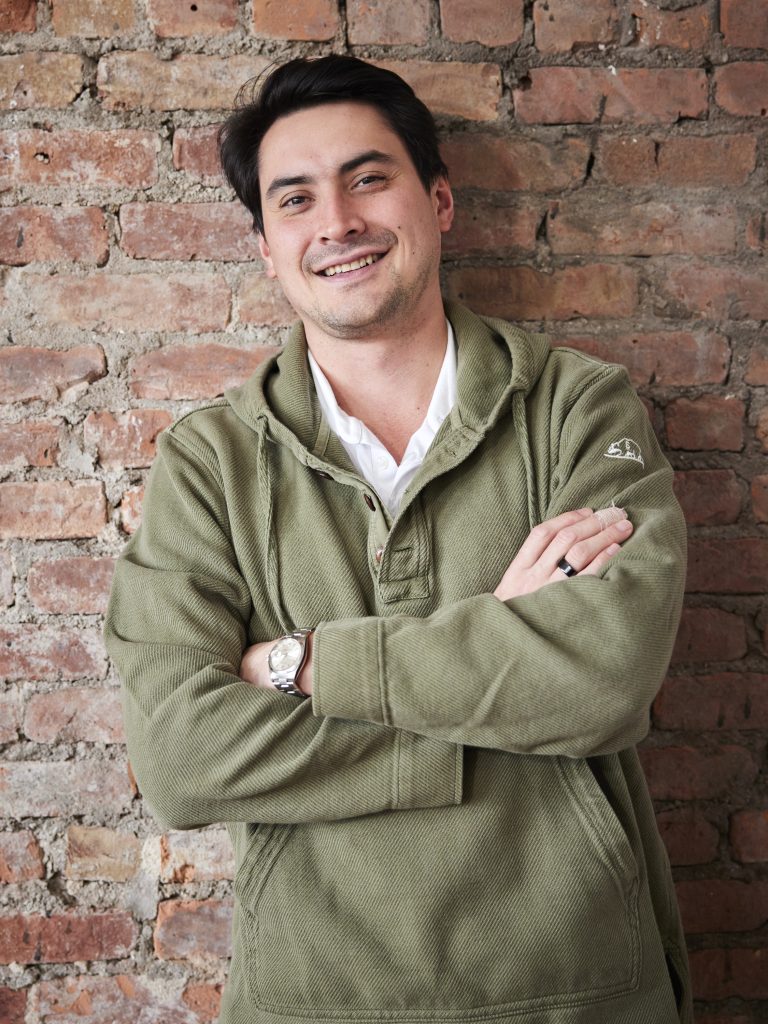A man wearing a green jacket standing in front of a brick wall with arms crossed.