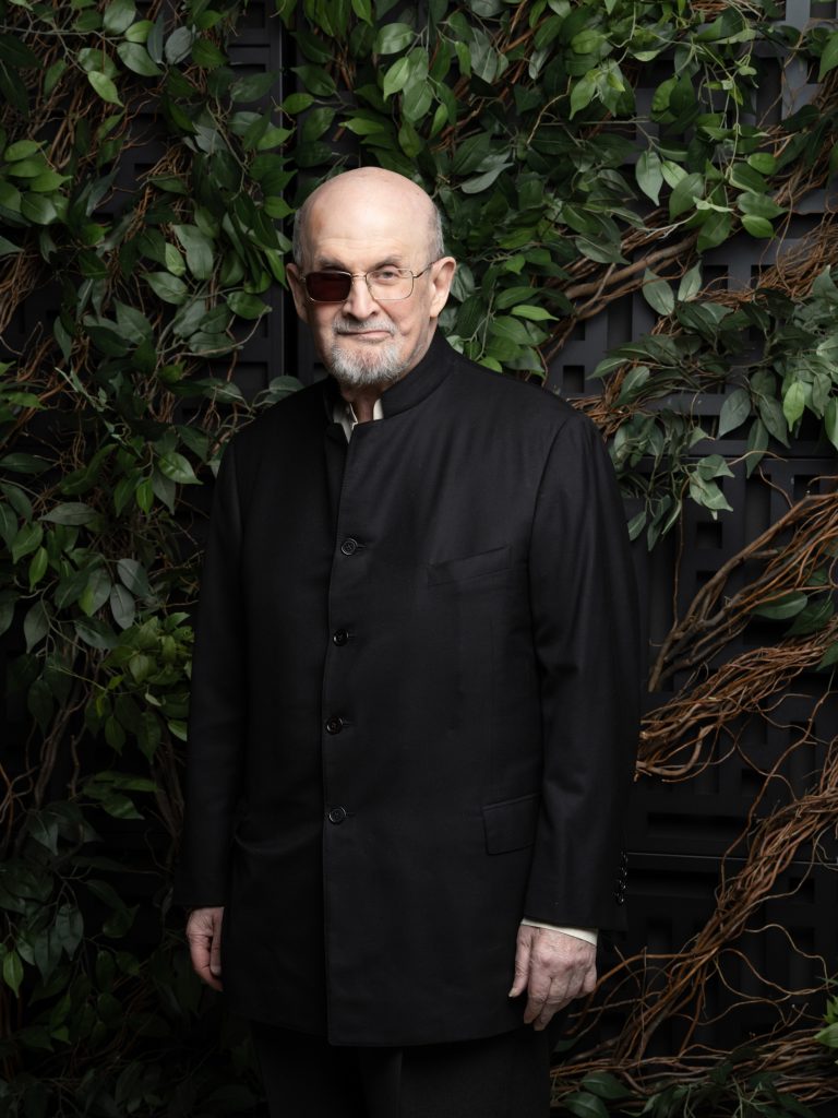 A man wearing dark clothing standing in front of foliage.