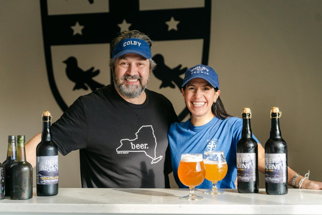 A man and a woman wearing Colby hats pose in front of a table of beer.