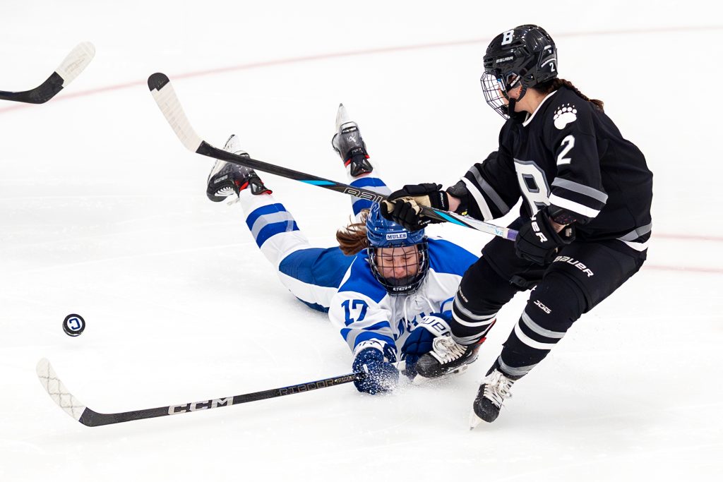 A hockey player diving on the ice as another player skates by.