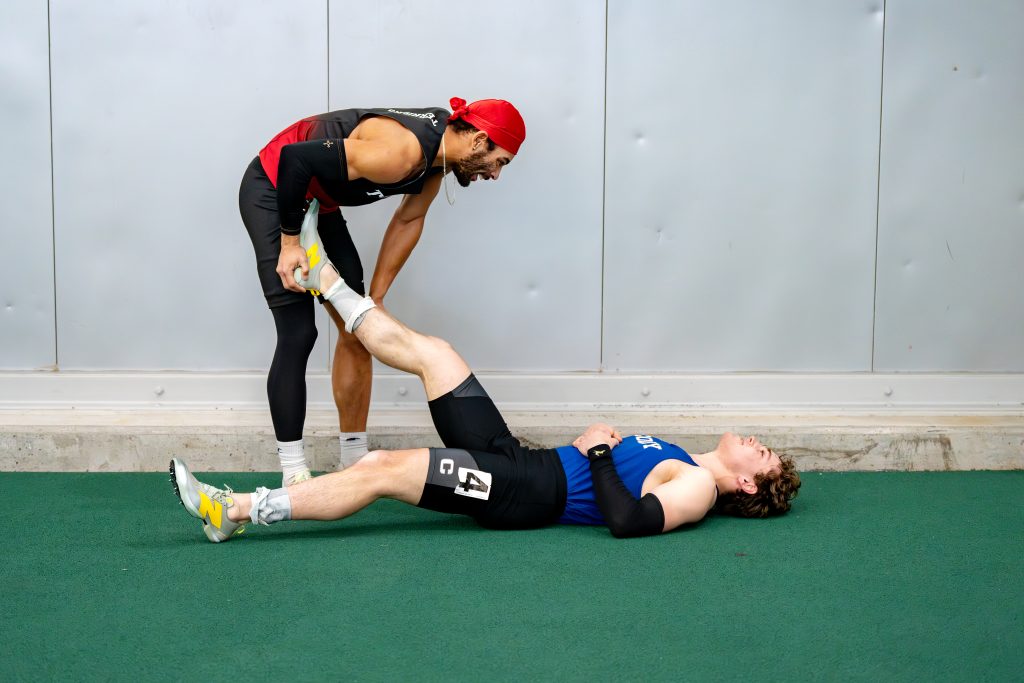 A track runner helps stretch the leg of another who is laying on the ground.