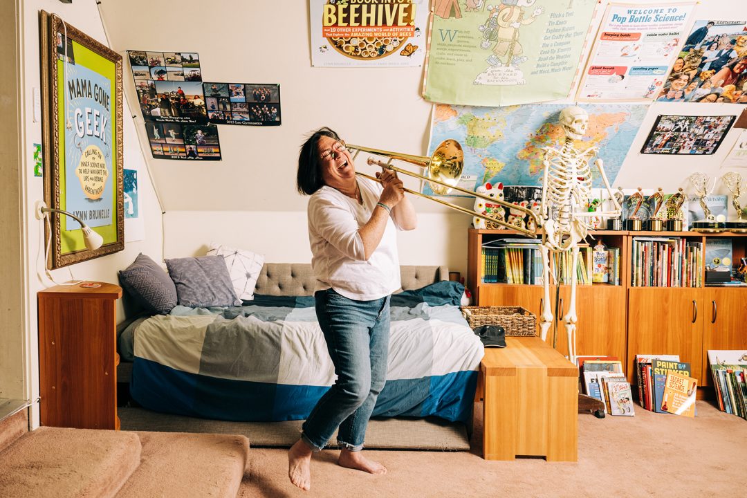 A barefoot woman plays the trombone in a creatively-decorated room.