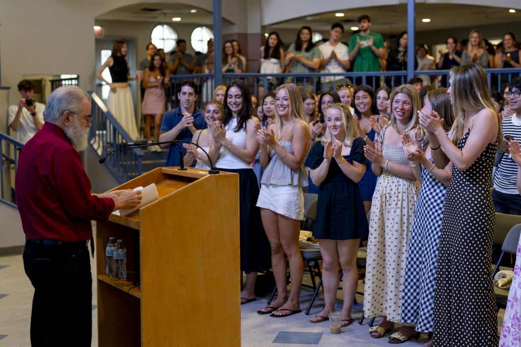 Students stand and cheer as a professor delivers remarks from a podium.