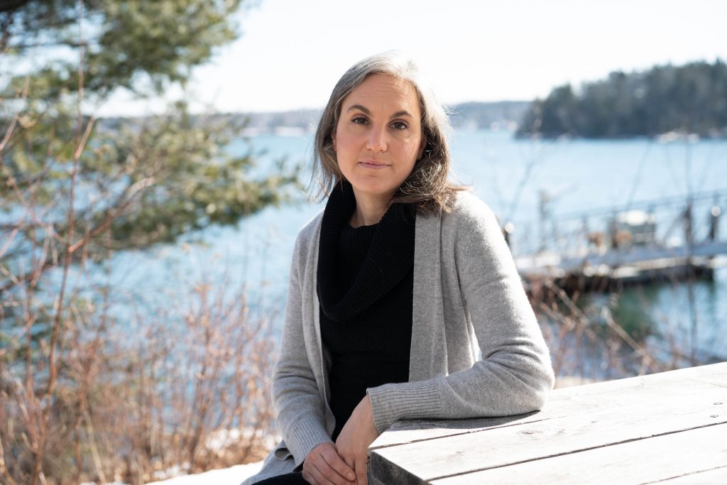 A woman sits at a picnic table with the ocean behind her. 