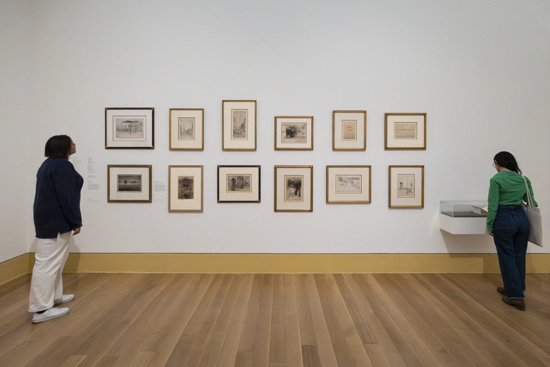 An image of paintings on the wall at a museum, with two people looking at them.