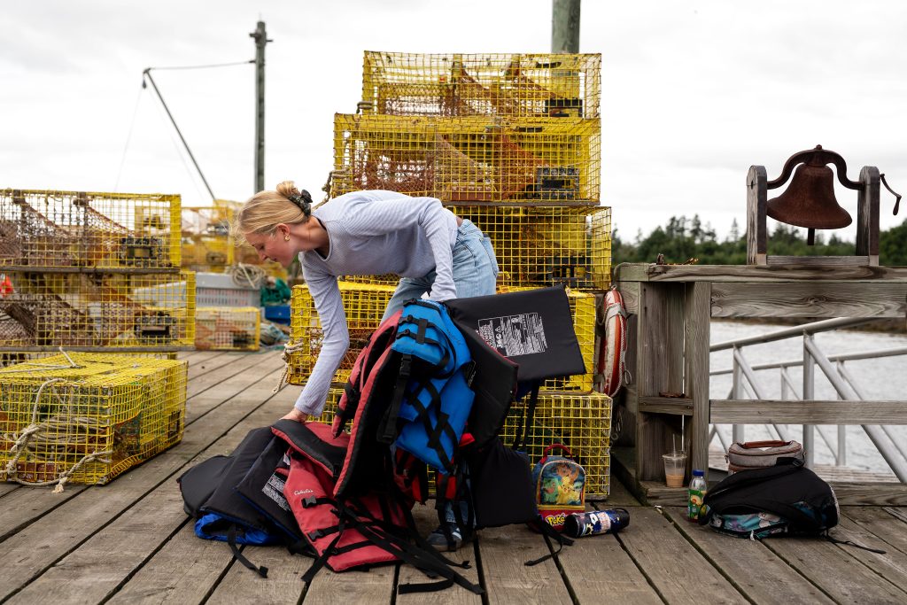 A person picks up a pile of life jackets with lobster traps piled up on the dock behind her. 