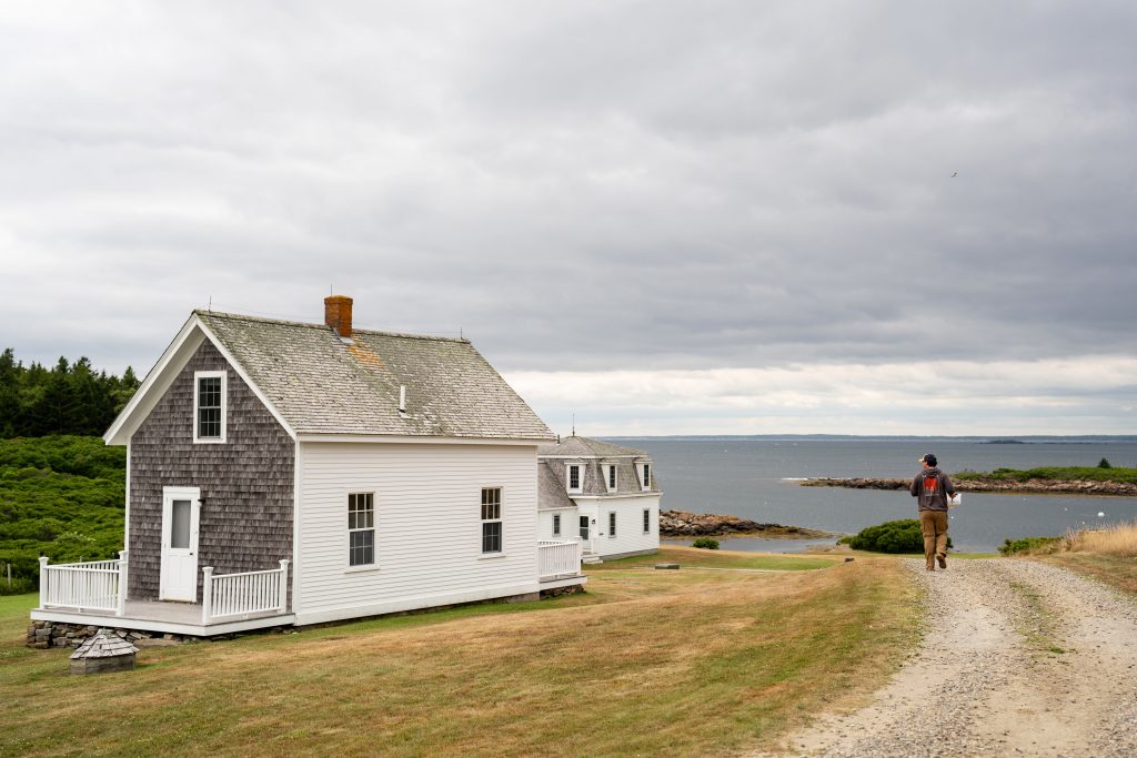 A person walks on a dirt road past some small houses. 