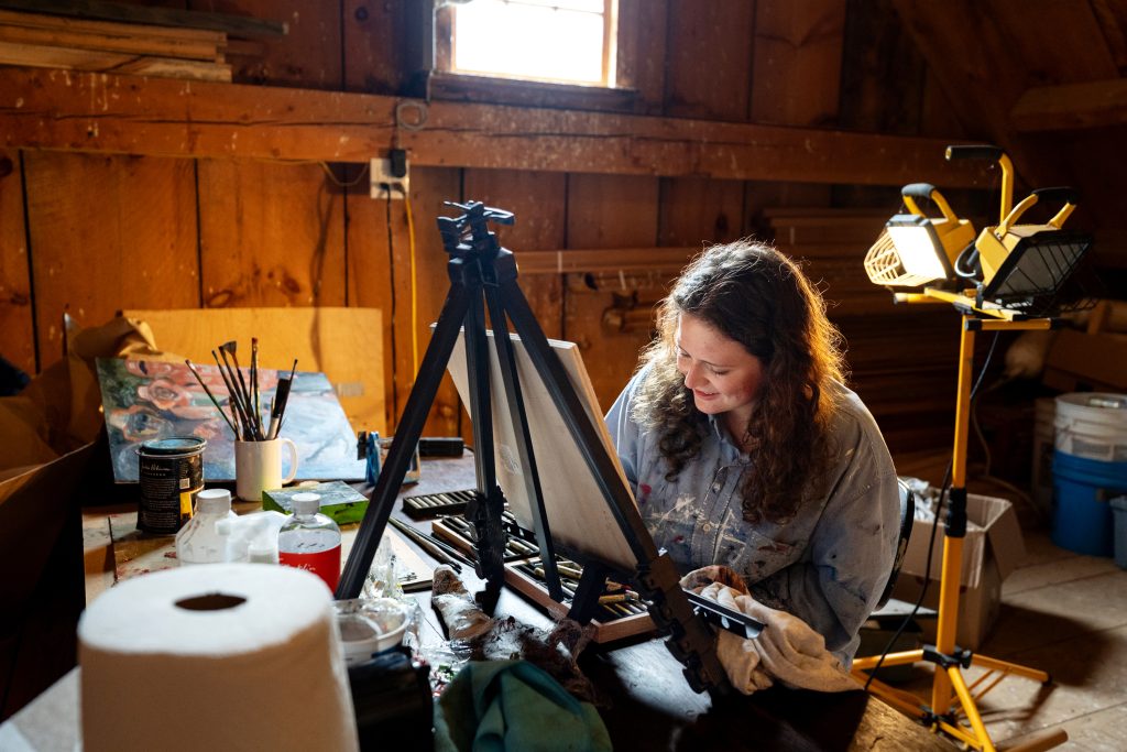 A woman works on a painting hanging on an easel in a rustic studio. 