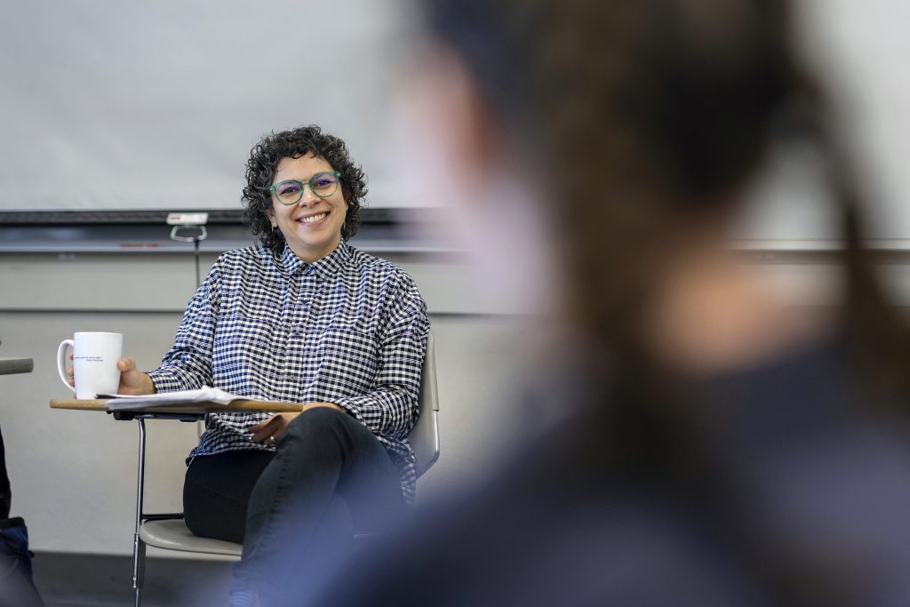 A smiling professor sits at a desk with a coffee cup. 