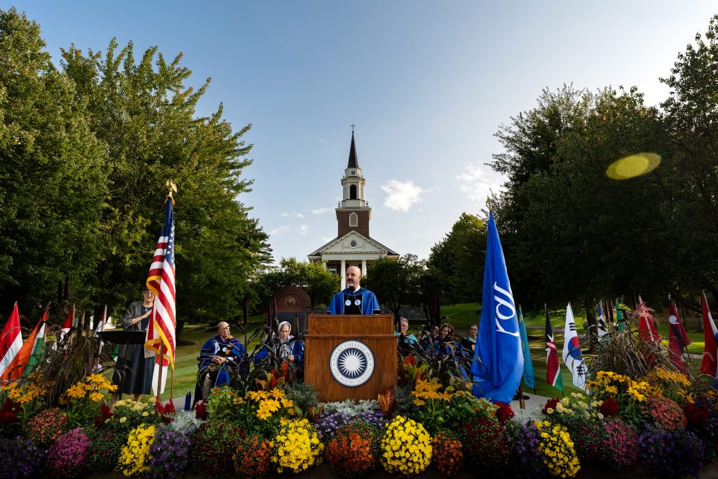 A man wearing an academic ceremonial gown delivers remarks at a podium set amid flowers on the Lorimer Chapel lawn. 