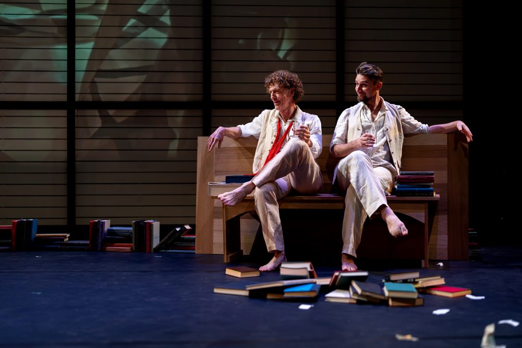 Two men in old-fashioned suits sit on a stage. 